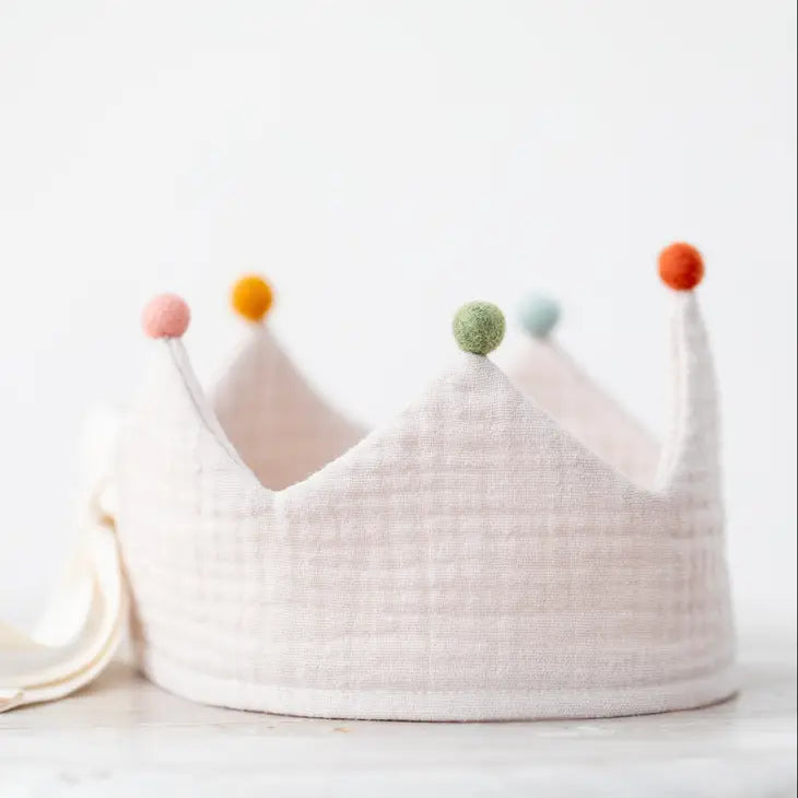 A soft pastel-colored Sand Reversible Birthday Crown with multicolored pom-poms attached to the points, set against a light, neutral background.