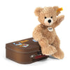 A Steiff, Fynn Teddy Bear in Suitcase with a Button in Ear, sitting on a brown suitcase adorned with travel stickers, waving its right paw, against a white background.