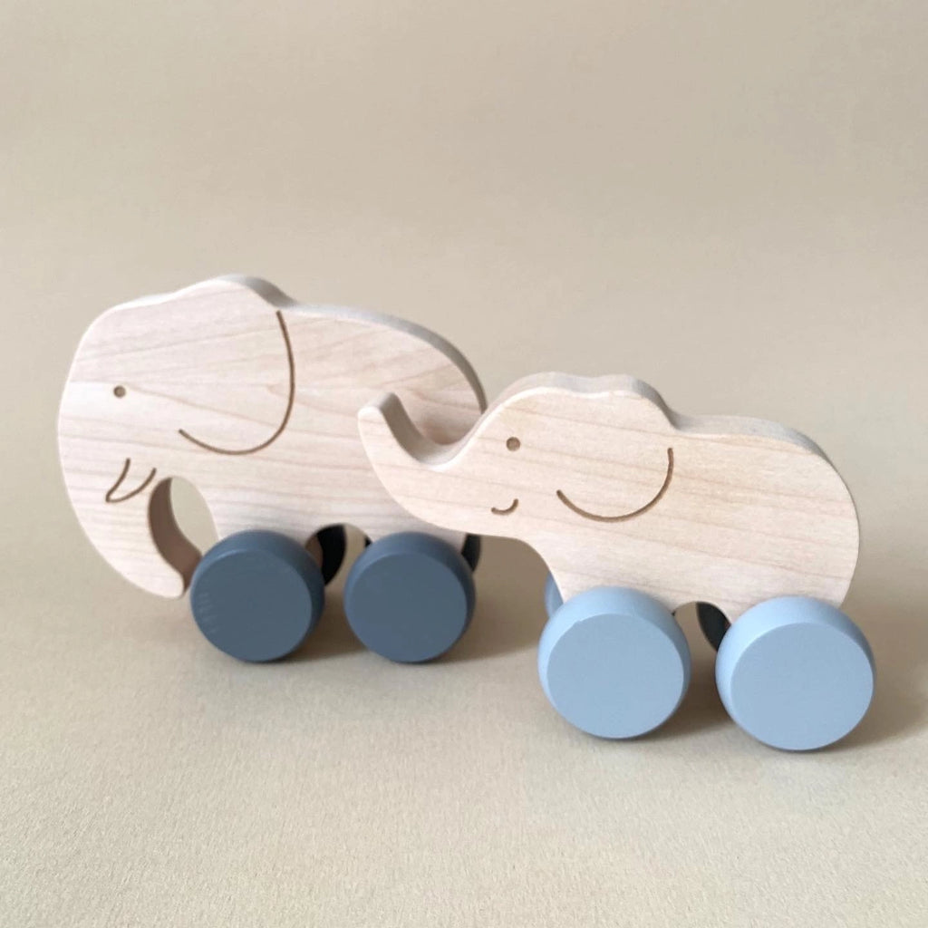 Two Wooden Mom and Baby Elephant Push Toy Set with light blue wheels on a beige background. The elephants, free of harmful chemicals, have simple, smiling features etched into the wood and are positioned side by side, with one being slightly larger than the other. Ideal for developing fine motor skills.