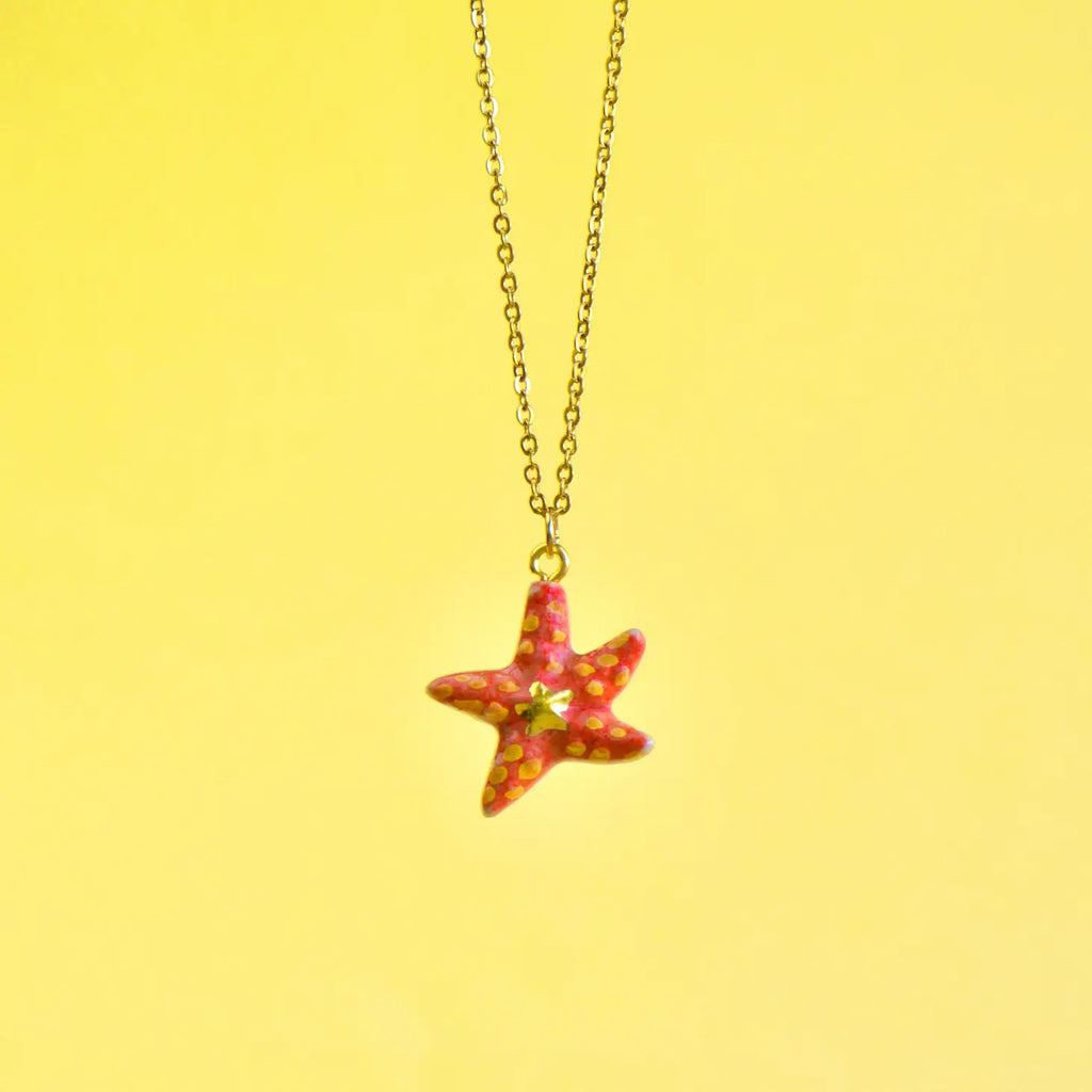 A Starfish Necklace with red speckles, hand painted and hanging from a delicate 24k gold plated steel chain against a soft yellow background.