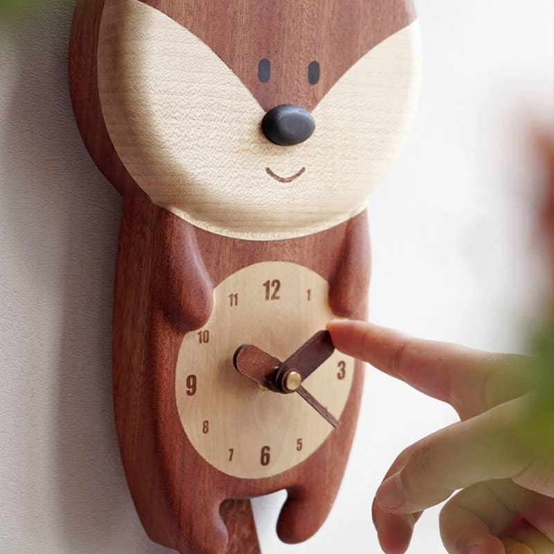 A hand points to the Handmade Wooden Fox Wall Clock, shaped like a fox with a round face, cheerful smile, and clear black numbers—an adorable addition to nursery decor.