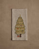 The Coral & Tusk Peek a Tree Tea Towel features an embroidered woodland Christmas tree with ornaments and gifts, set on a beige linen cloth against a textured tan background.
