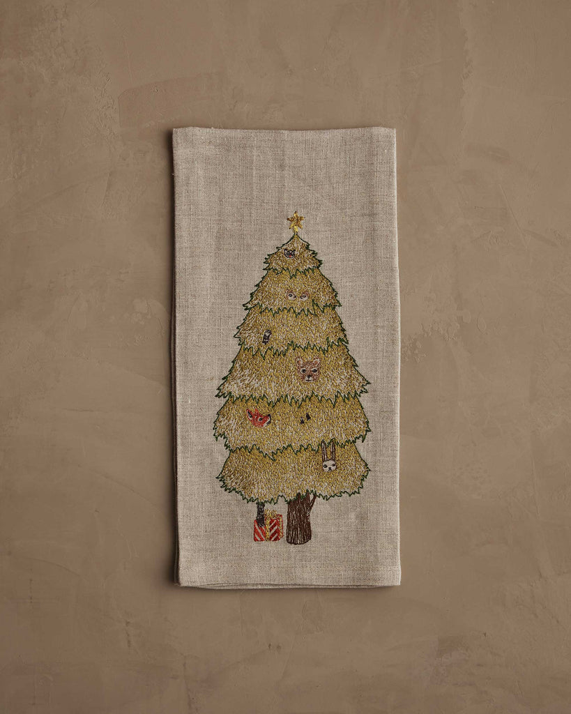 The Coral & Tusk Peek a Tree Tea Towel features an embroidered woodland Christmas tree with ornaments and gifts, set on a beige linen cloth against a textured tan background.