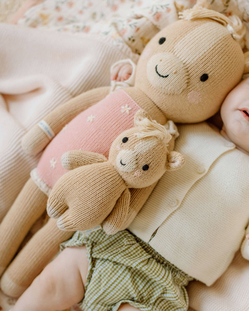 A baby lies next to two hand-knit Cuddle + Kind Sophia the Pony dolls, one large and one small. The baby wears green gingham shorts and a cream cardigan. These soft, fair trade ponies feature gentle smiles and light brown yarn.