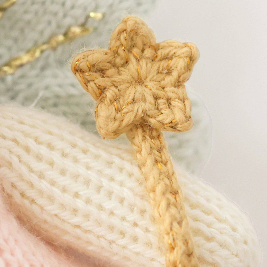 A close-up of Cuddle + Kind Tiny Lily the Fairy’s beige knitted star, attached to a braided cord and resting on cream and pastel pink knits—an ideal sustainable, fair trade accessory for this hand-knit doll.