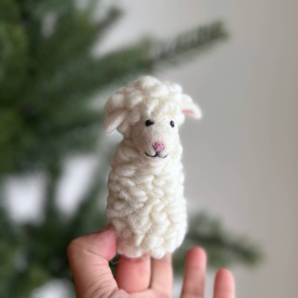 A hand holds the Felt Finger Puppet: Fluffy Sheep - White, a small handmade wool sheep with a curly body and embroidered face—perfect for imaginative play or storytelling—set against a blurred green background.