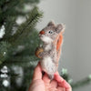 A hand holds the Felt Finger Puppet: Gray Squirrel—an eco-friendly wool toy with an acorn, set near pine branches. Perfect as a woodland gift for kids or handmade toy lovers. The softly blurred background highlights its charming detail.