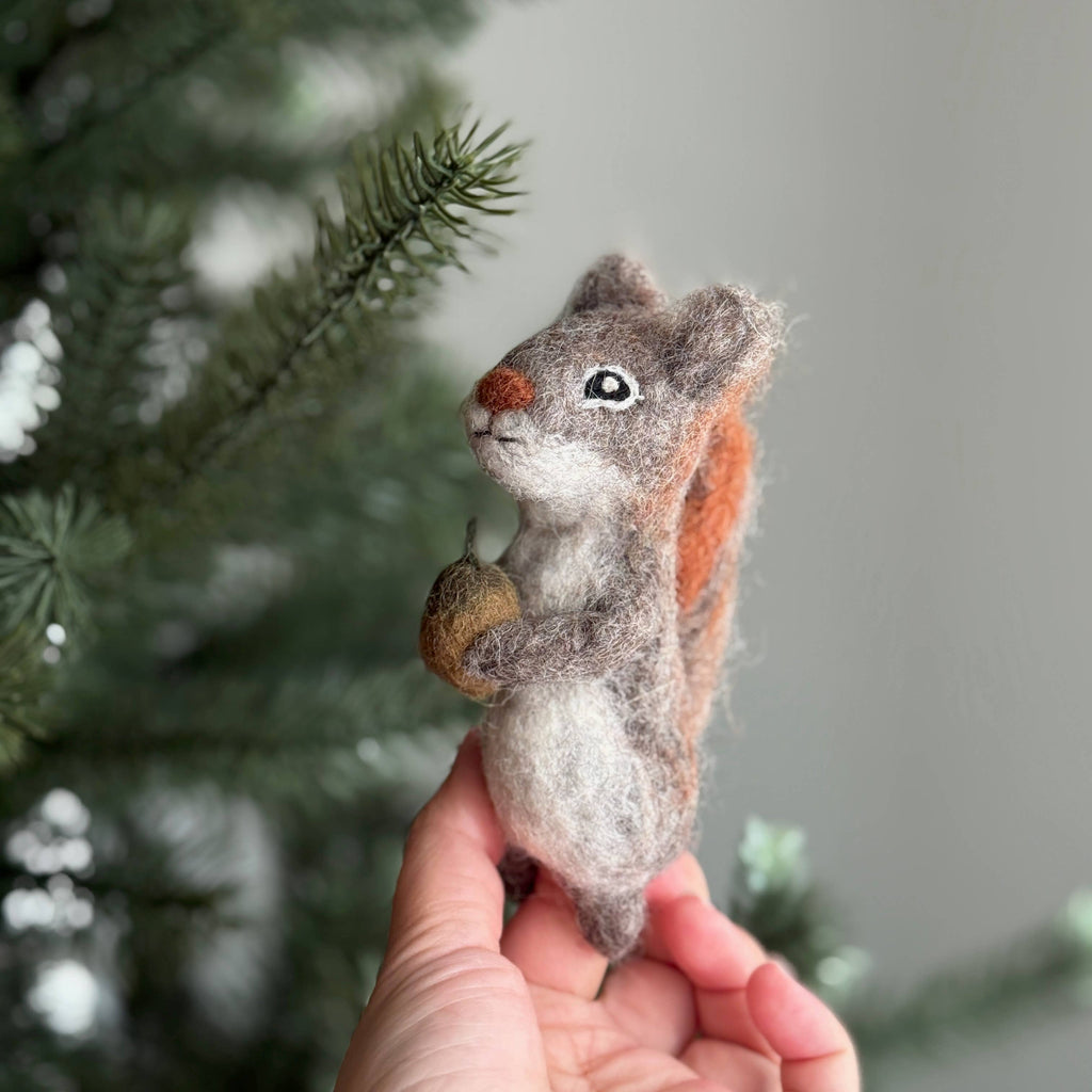 A hand holds the Felt Finger Puppet: Gray Squirrel—an eco-friendly wool toy with an acorn, set near pine branches. Perfect as a woodland gift for kids or handmade toy lovers. The softly blurred background highlights its charming detail.