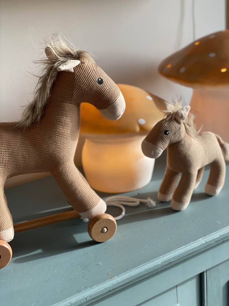 Two brown plush toy horses, one of which is Pull-Along Henry—a delightful wooden horse toy on wheels with a pull string—stand on a blue-green surface. A mushroom-shaped lamp from Egmont Toys glows softly in the background, adding a gentle warmth to the scene.
