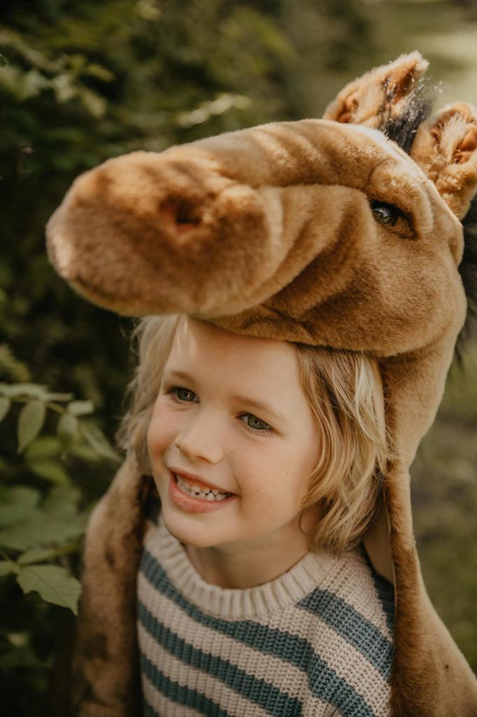 A smiling blonde-haired child stands outdoors in natural light near green foliage, wearing a striped sweater and the Costume - Brown Horse, a large plush horse hat that resembles an adorable horse disguise.