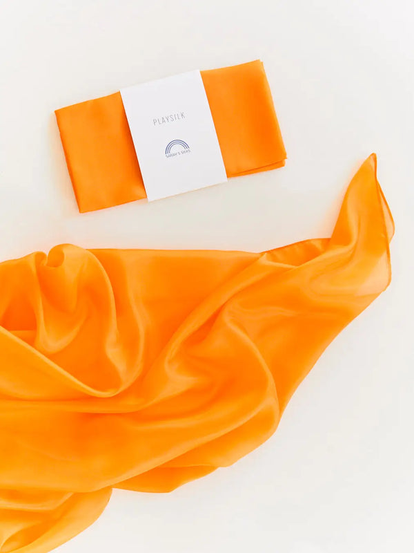 A vibrant orange Sarah's Silk Playsilk elegantly draped next to an orange packaging box with the word "pleasure" and a small rainbow-like logo on it, all against a white background.