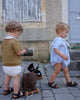 Two toddlers in vintage-style outfits pull Pull-Along Walter toys with wooden wheels along a cobblestone street, developing their gross motor skills. One wears a brown sweater, the other a blue romper, with weathered building doors visible in the background.