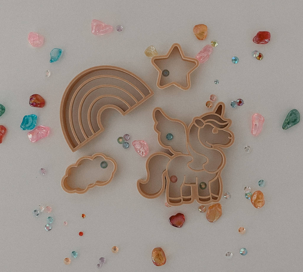The Unicorn Cutter Set - Eco Playdough Tool features tan plastic cookie cutters in whimsical shapes including a rainbow, star, cloud, and unicorn. Surrounded by vibrant gemstones on a light background, these eco-friendly sensory play tools inspire imaginative creativity with the unique unicorn cutter included in the set.