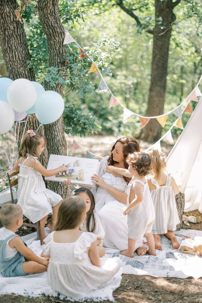 A woman reads Winnie-the-Pooh: Finding Feelings to children outdoors on a picnic blanket, surrounded by trees, balloons, and bunting for a fun woodland storytime.