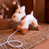 Pull-Along Lola is a fluffy white stuffed dog with a red plaid collar, attached to wooden wheels and a pull string—ideal for toddlers as a gift or for developmental play.
