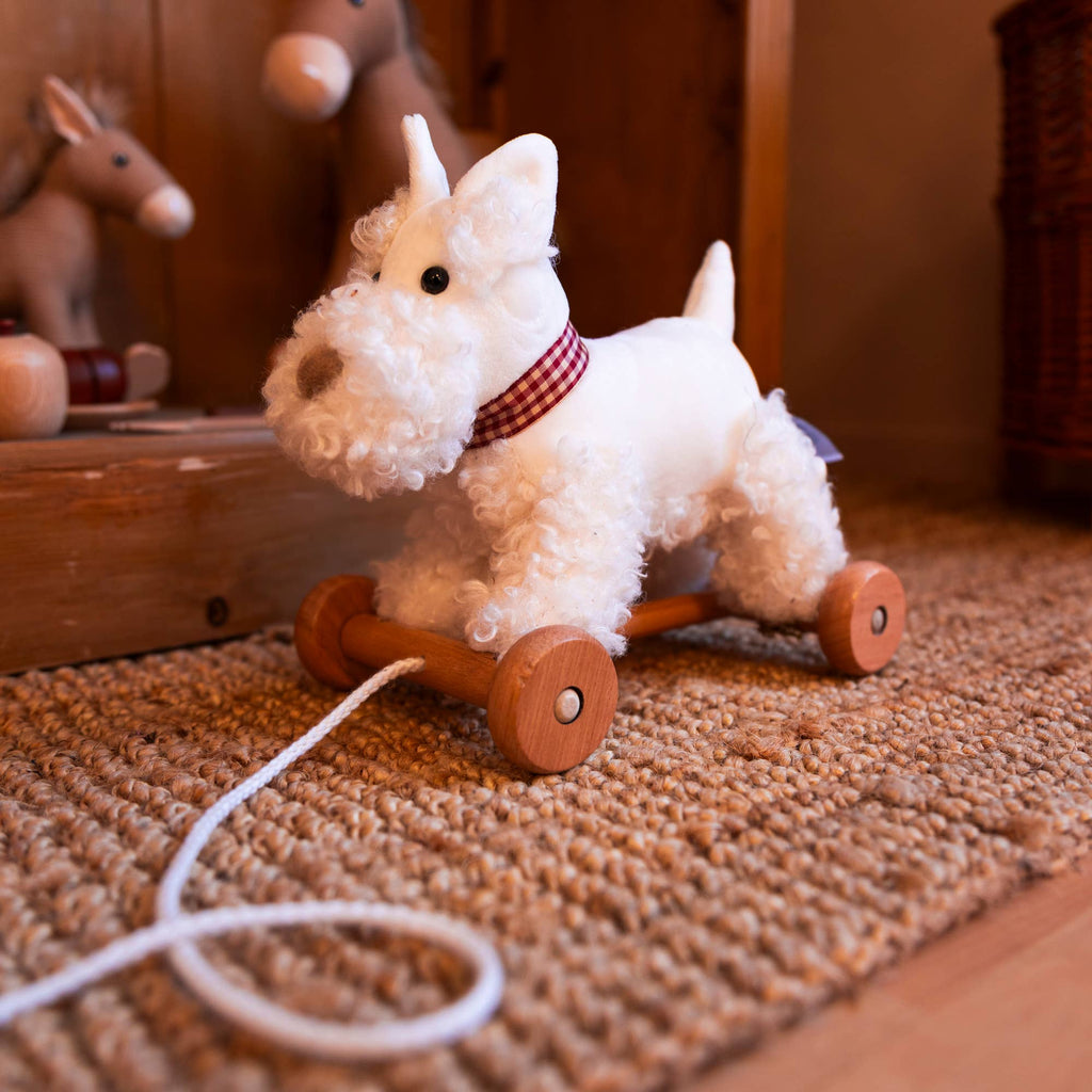 Pull-Along Lola is a fluffy white stuffed dog with a red plaid collar, attached to wooden wheels and a pull string—ideal for toddlers as a gift or for developmental play.