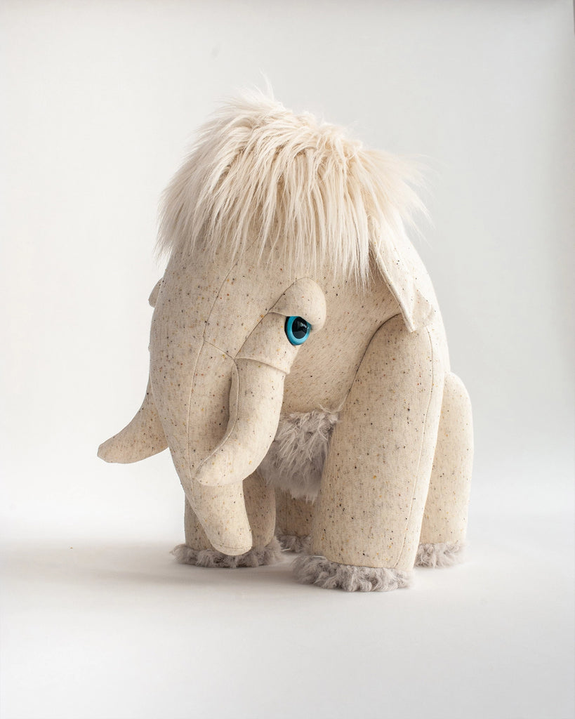 A BigStuffed Albino Mammoth Stuffed Animal with light beige textured fabric, fluffy fur on its head and ankles, and large blue eyes stands against a plain white background.