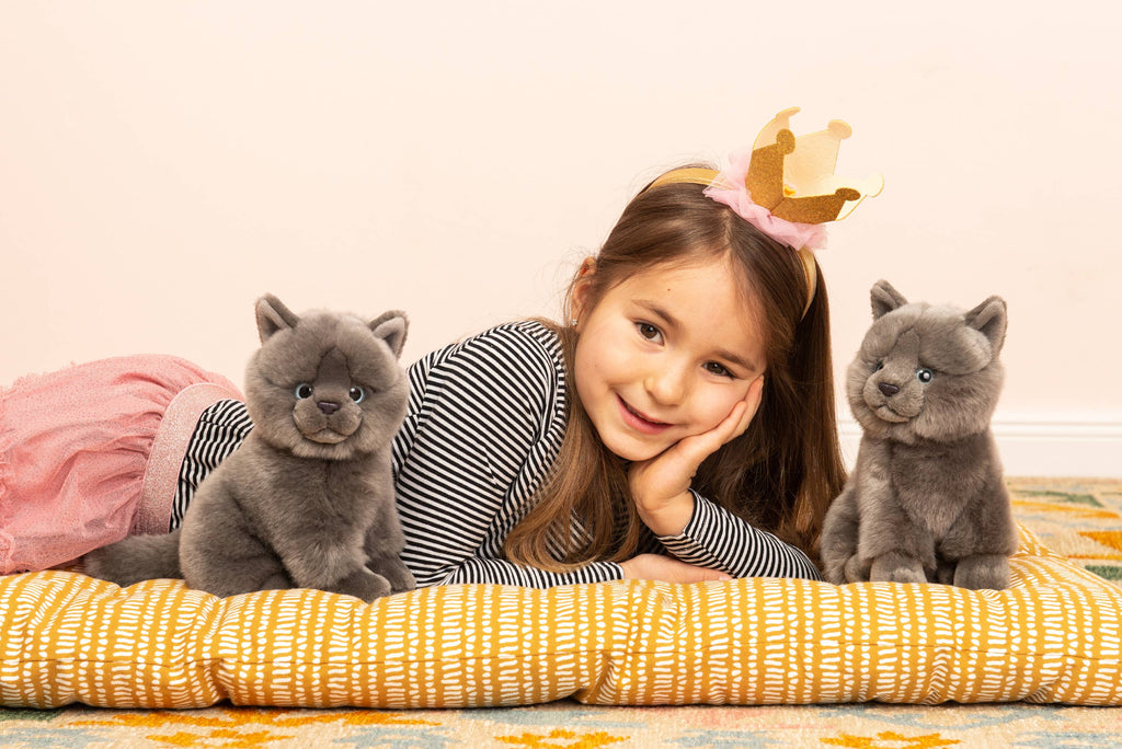 A young girl wearing a gold paper crown lies on her stomach, smiling. She rests her head on her hand and lies on a cushioned surface with two Teddy Hermann Gray Sitting Cat Stuffed Animals beside her. She wears a striped shirt and pink skirt, and the background features a soft, neutral-toned wall.