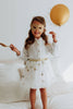 A young girl stands on a bed wearing a Pink Sequins Mask—an enchanting costume accessory—and a white dress with silver stars. Holding a gold balloon and decorative spoon, she looks cheerful and playful, ready for glamorous evenings of fun.