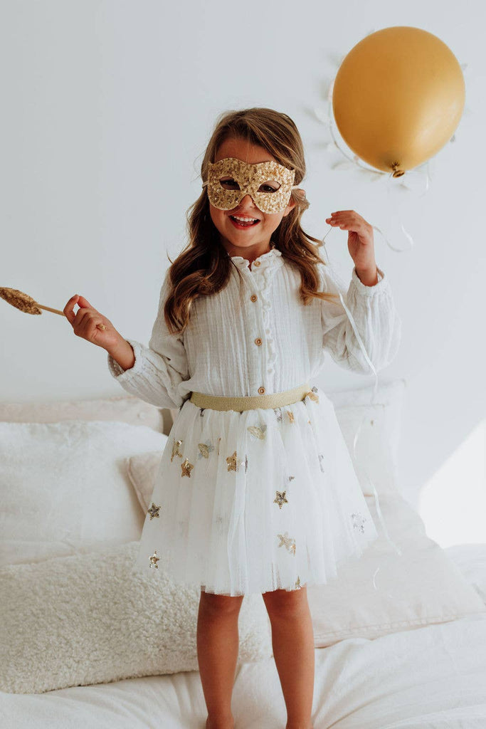 A young girl stands on a bed wearing a Pink Sequins Mask—an enchanting costume accessory—and a white dress with silver stars. Holding a gold balloon and decorative spoon, she looks cheerful and playful, ready for glamorous evenings of fun.