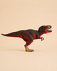 The Schleich Red Tyrannosaurus Rex Dinosaur Toy, featuring red and black coloring, stands on two legs against a plain beige background.