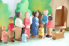 A group of colorful, handcrafted wooden figurines, including men, women, and children, stand together on a green surface with trees in the background. The group appears to be gathered around an Ostheimer Child In Cradle. This picturesque scene of Ostheimer wooden toys invites imaginative play in a quaint, natural environment.