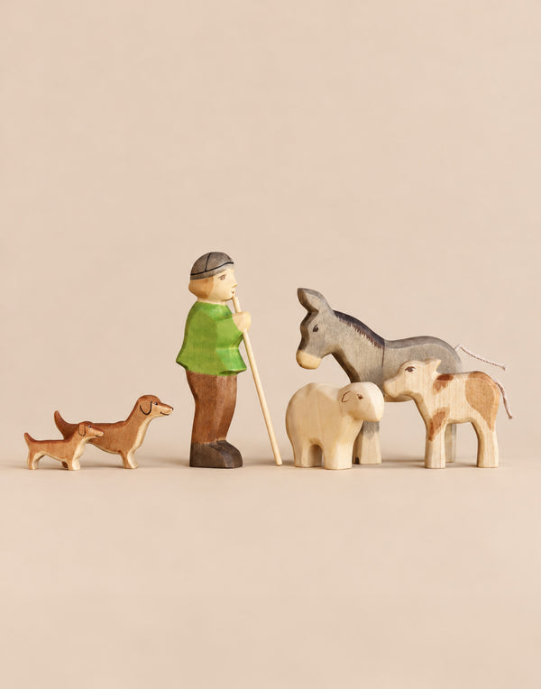 This Handmade Holzwald Shepherd & Farm Animals Set features a shepherd with a staff, two dachshunds, a donkey, a sheep, and a calf, all hand-carved from wood.