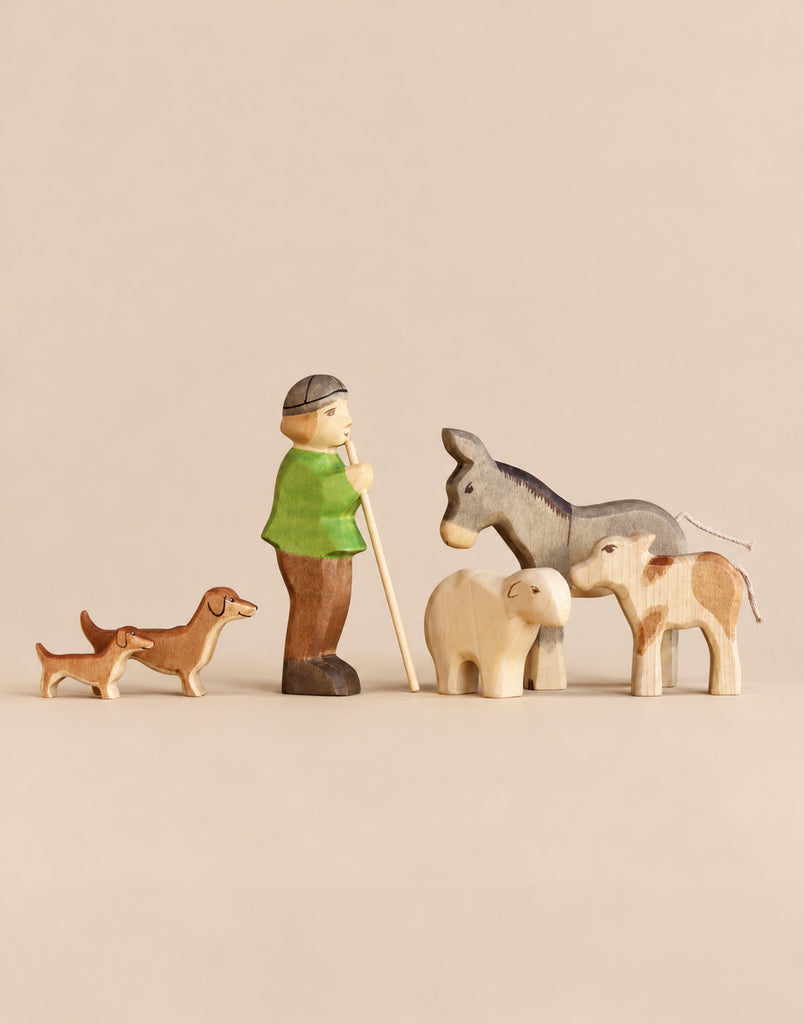 This Handmade Holzwald Shepherd & Farm Animals Set features a shepherd with a staff, two dachshunds, a donkey, a sheep, and a calf, all hand-carved from wood.