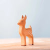 The Bumbu Handmade Wooden Fawn, crafted from sustainably sourced beechwood and featuring a light brown finish with white spots, stands on a smooth wooden surface set against a soft pale blue background.