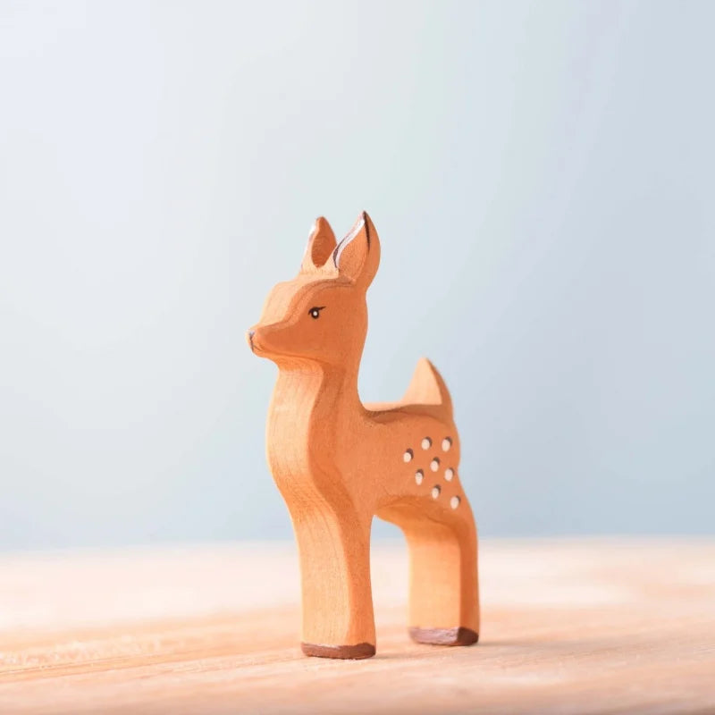 The Bumbu Handmade Wooden Fawn, crafted from sustainably sourced beechwood and featuring a light brown finish with white spots, stands on a smooth wooden surface set against a soft pale blue background.
