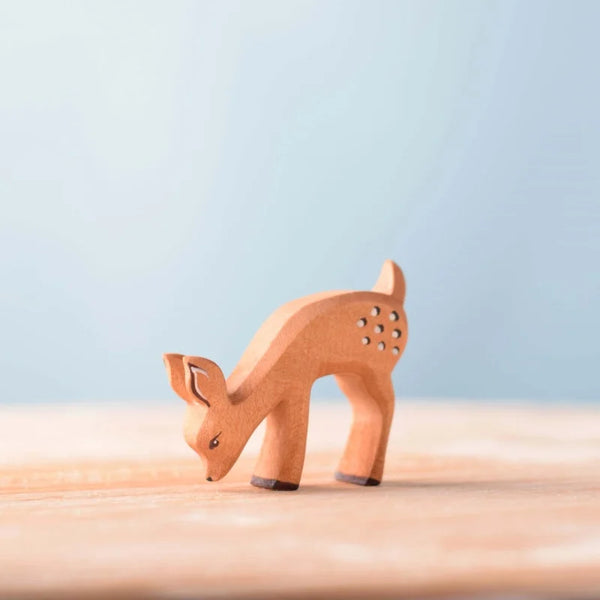 The Bumbu Handmade Wooden Fawn Eating is a charming, eco-friendly wooden toy with white spots, handcrafted for imaginative play, shown grazing on a light surface against a soft blue background.