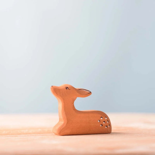 The Bumbu Handmade Wooden Fawn Resting, a small brown handcrafted wooden figurine with simple painted features, sits on a light wood surface against a pale blue background.