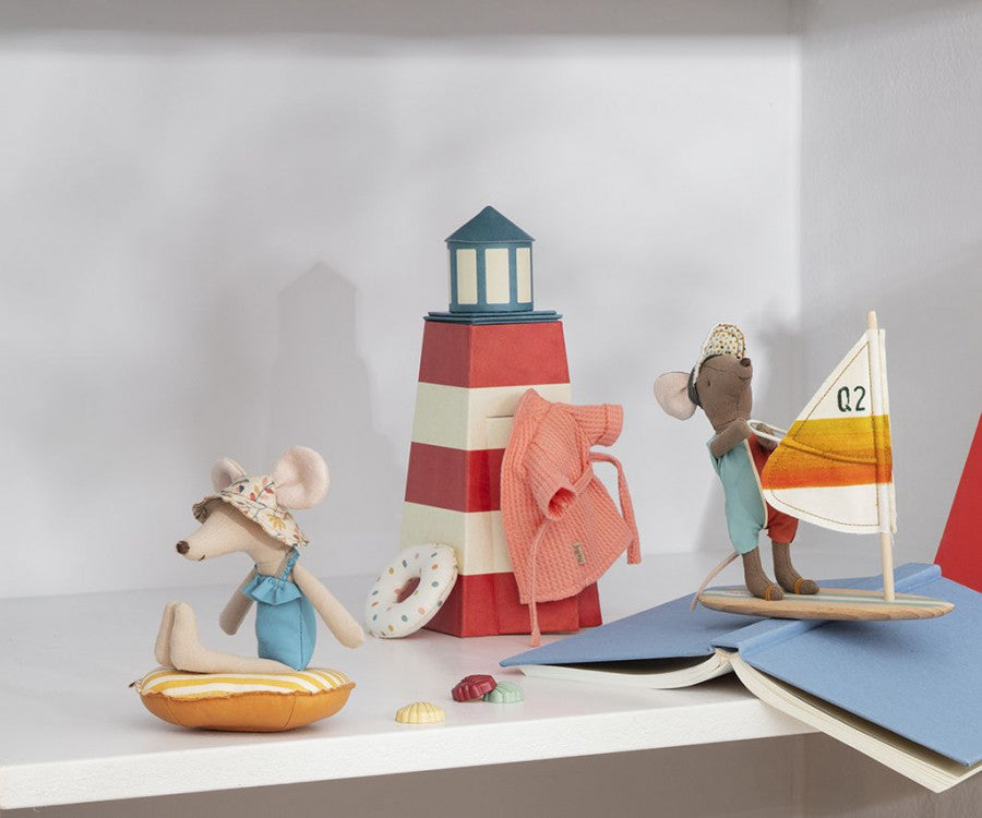 Two toy mice, dressed in summer attire, enjoy a beach-themed setup. One mouse, cozy in a Maileg Extra Clothing: Bathrobe - Coral, sits on a yellow float while the other, wearing a polka-dot hat, stands on a sailboat with a striped sail. A red and white striped lighthouse prop and a life ring enhance the scene.