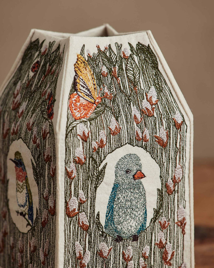 A fabric lampshade with embroidered birds, flowers, and a butterfly in earthy tones reflects the charm of the Coral & Tusk Birdhouse Tissue Box Cover, featuring detailed nature scenes on every panel.