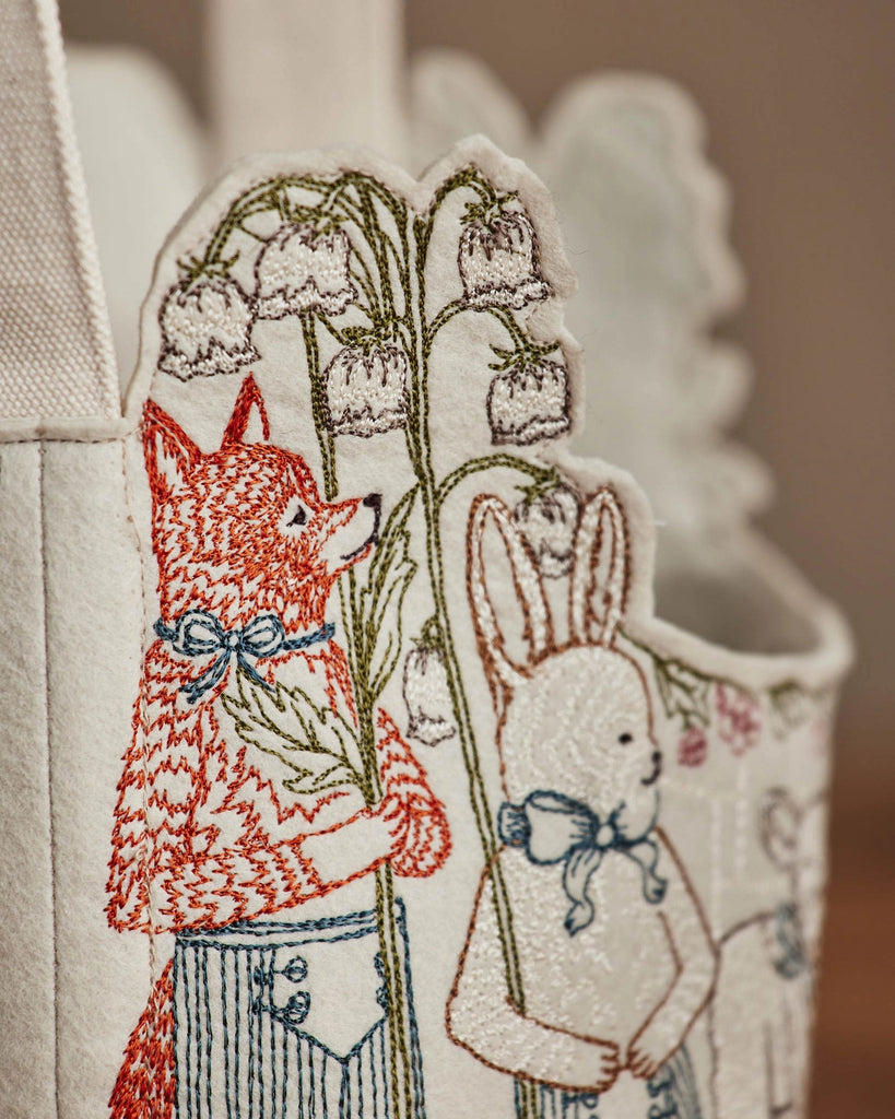 Close-up of embroidered fabric from the Coral & Tusk Easter Parade Basket, showing a red fox and white rabbit in clothes holding lily of the valley sprigs, set against a softly blurred background for added whimsy.