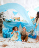 Three children are joyfully playing in front of a festive table adorned with blue and white fabrics. One child is creatively using Sarah's Silk Giant Snow Playsilk, holding up the blue cloth as others look on, amidst faux snow and snowflake decorations, fueling open-ended play in a winter wonderland scene.