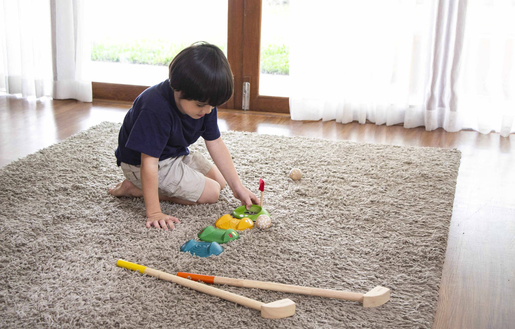 A young child plays indoors on a beige carpet with the Mini Golf Set, using colorful crocodile pieces, small balls, and two wooden mallets with colored handles as sunlight streams through large windows in the background.