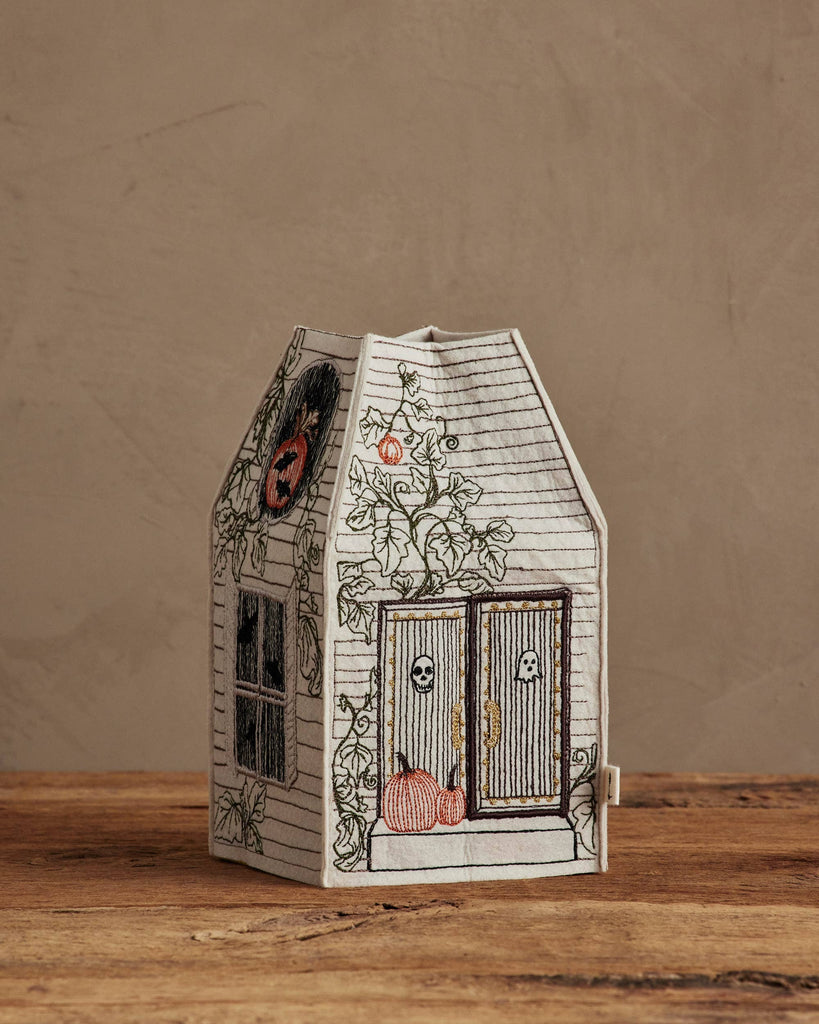 The Coral & Tusk Haunted House Tissue Box Cover, shaped like a house with embroidered pumpkins, skulls, and vines, sits on a wooden surface against a beige wall.