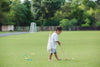 A young child in shorts and a t-shirt plays barefoot on grass with the Mini Golf Set, swinging a wooden stick at a ball. The child carries a bag over one shoulder, with trees and a soccer goal in the background.