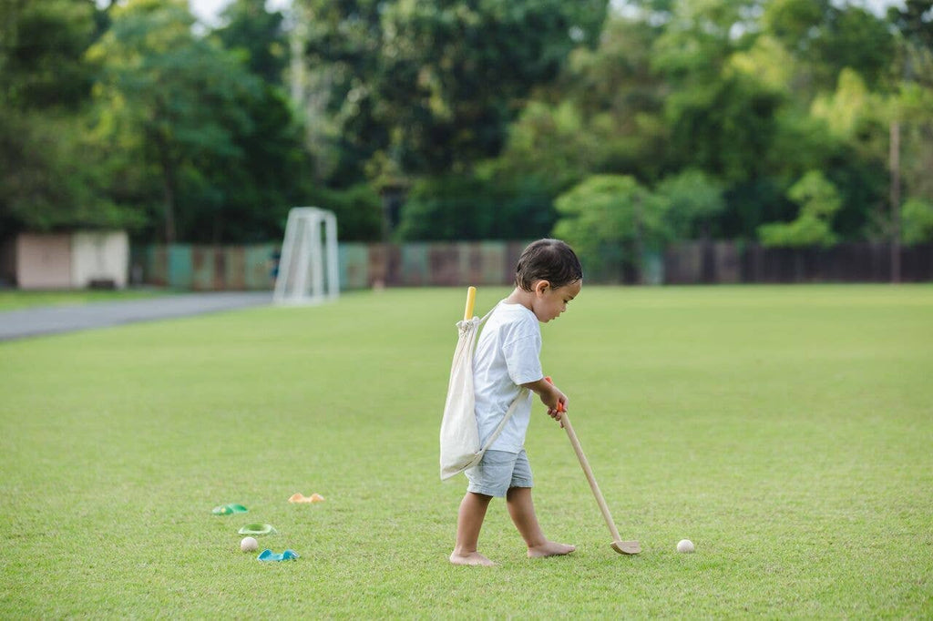 A young child in shorts and a t-shirt plays barefoot on grass with the Mini Golf Set, swinging a wooden stick at a ball. The child carries a bag over one shoulder, with trees and a soccer goal in the background.