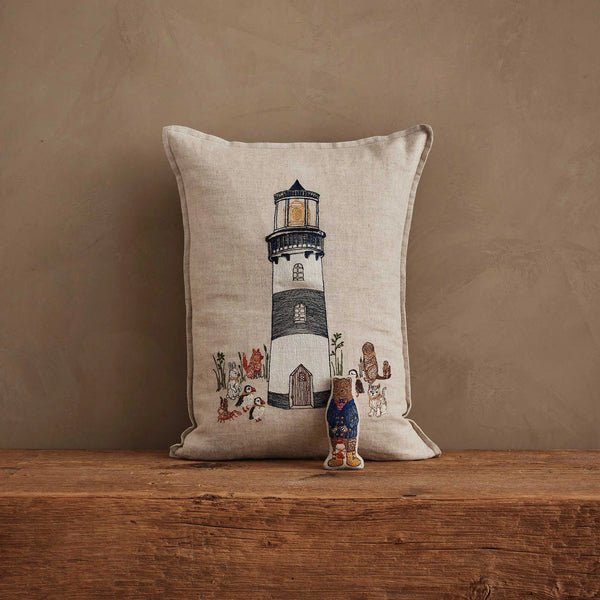 The Coral & Tusk Lighthouse Friends Pocket Pillow, featuring embroidered linen with a lighthouse and animals, stands against a brown textured wall with a decorative doll in front. Its cover meets Oeko-Tex Standard 100 safety standards.