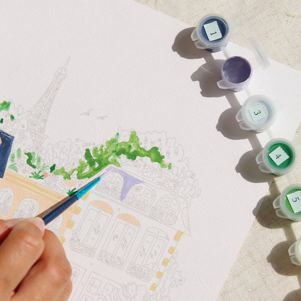An aspiring artist uses the Paint By Number Kit - Paris, applying green paint to a scene of the Eiffel Tower, with numbered paint pots close by, sparking creativity.