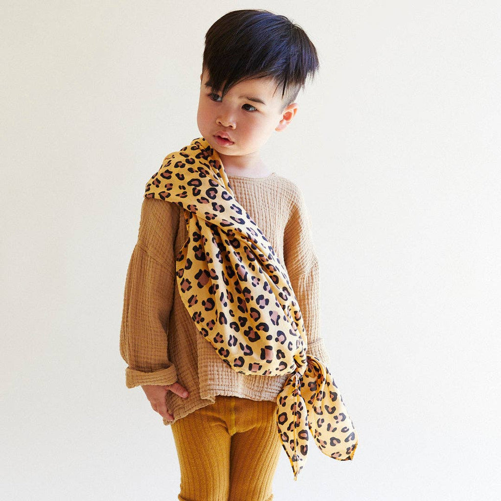 A young child with short dark hair stands against a plain light background, wearing a beige textured sweater, mustard ribbed pants, and draping Sarah's Silks Animal Print Playsilk - Cheetah over one shoulder.