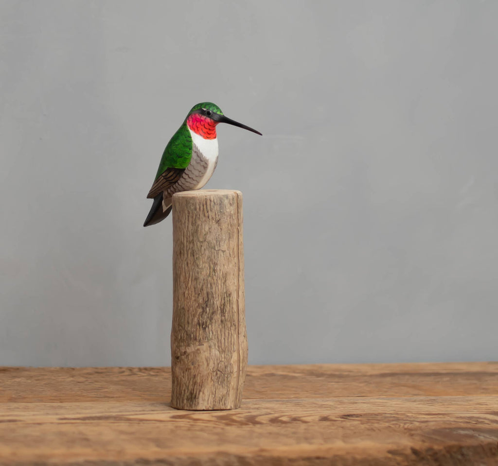 The Hand Carved Ornament - Humming Bird - 7.5"H features a painted hummingbird with iridescent green feathers and a red throat perched on a wooden log, set against a plain gray background and wooden surface.