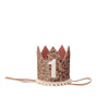 A small, glittery **Rose Gold Glitter Crown** with a felt texture, bearing a white number "1" on the front. Perfect for birthday celebrations, the crown has a pink interior lining, pom-pom trim at the base, and two attached strings on either side. It is set against a plain white background.