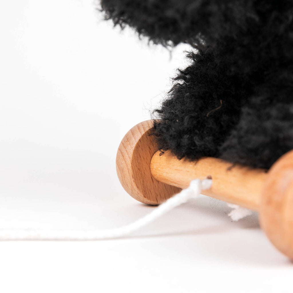 A close-up of a wooden wheel attached to Pull-Along Walter, a fluffy black pull-along toy, on a white background with a white pull string visible in the foreground.