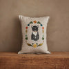 The Coral & Tusk Kitten Cameo Pillow in beige showcases an embroidered black and white kitten cameo, accented by vibrant spring flowers and leaves, displayed on a wooden surface against a plain textured wall.