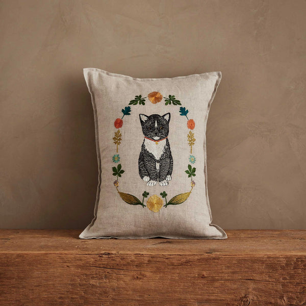 The Coral & Tusk Kitten Cameo Pillow in beige showcases an embroidered black and white kitten cameo, accented by vibrant spring flowers and leaves, displayed on a wooden surface against a plain textured wall.