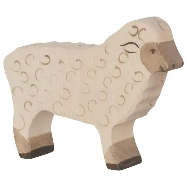 A carved wooden sheep, part of the **Holztiger Sheep, Standing** collection, stands on a flat surface. Made in Europe, the sheep is light in color with darker shades on its hooves and snout. The body is detailed with small swirls to represent fleece, and the face has a calm expression with closed eyes.