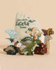 A set of colorful dinosaurs from the Easter Basket Gift Set is stacked in front of a drawstring bag labeled “Dinosaurs Stacker Tower.” Both the bag and dinosaurs display earthy tones and painted skeletal accents.